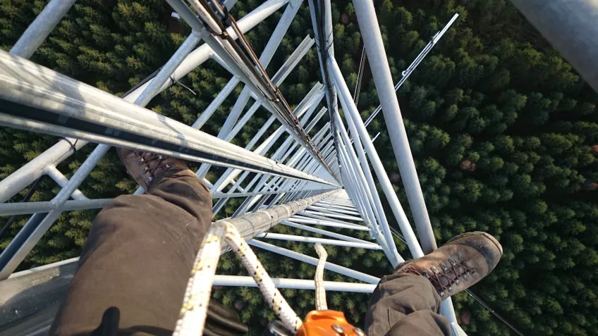 Legs and feet of a person sitting high up in a measuring station. Photo taken from above, showing trees far below.