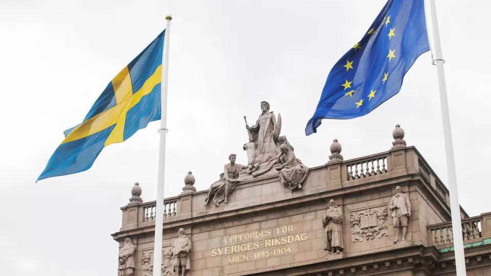 En del av riksdagshuset samt Sveriges och EUs flaggor. Foto.