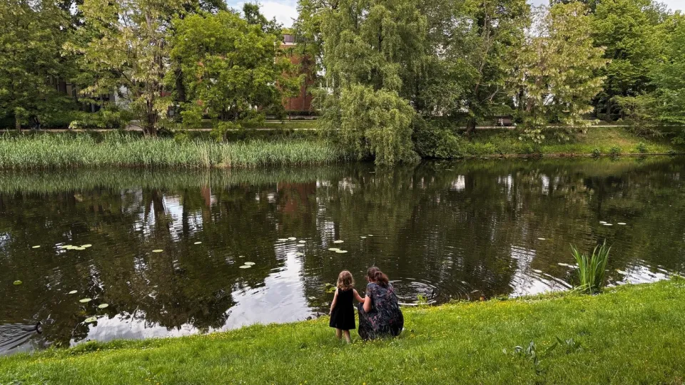 Kvinna och barn står på gräsmatta och tittar på en damm i parkmiljö.