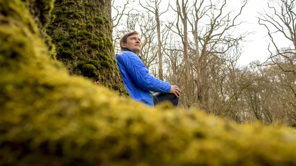 Yann Clough sitter med ryggen mot ett träd i en skog. Foto.